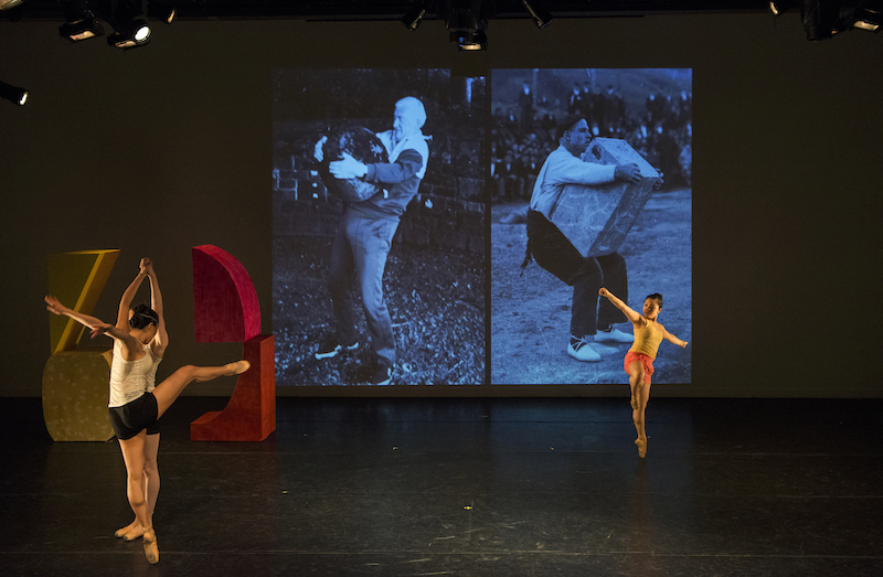 Three women dance on pointe as a black and white film projects behind them and two brightly colored larger-than-life shapes sit upstage